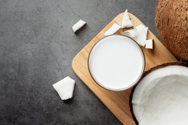 Fotografia de um vidro com óleo de coco branco e cremoso, colocado sobre uma tábua de madeira. Ao redor do copo, há pedaços de coco fresco, além de uma metade de coco com sua polpa branca exposta. O fundo é um tom escuro de cinza, criando um contraste com os elementos brancos.